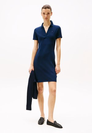 Marineblå polo kjole med korte ærmer, V-hals og tætsiddende silhuet. Accessories inkluderer en vævet marineblå cardigan og sorte loafers.
