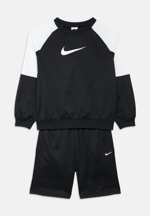 Schwarzes Sportset bestehend aus einem langärmeligen Oberteil mit weißen Schulterakzenten und weißem Nike-Logo, kombiniert mit schwarzen Shorts, beide aus weichem Stoff gefertigt.
