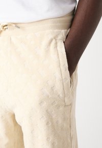 Cream-colored terry joggers with a textured pattern, elastic waist with drawstring, and side pockets. Smooth fabric with a soft finish.