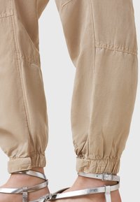 Béžové kalhoty s elastickými manžetami vyrobené z hladké a lehké tkaniny. Mají postranní kapsy a mírně nařasené kotníky. S kombinací stříbrných sandálů.
