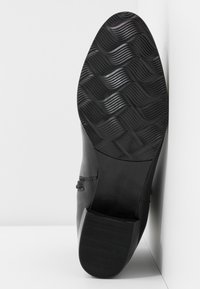 Semelle de botte noire avec motif de bande de roulement ondulé texturé et talon lisse, positionnée verticalement contre une surface d'angle blanche.
