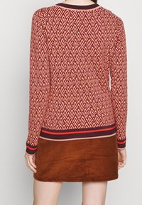 Rot- und cremefarbener Pullover mit geometrischem Muster, gerippten Bündchen und einem Halsausschnitt, kombiniert mit einem braunen Cordrock.