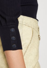 Marineblauer gerippter Pullover mit drei dunklen Knöpfen am Bündchen, kombiniert mit blassgelben, strukturierten Hosen mit rissiger Oberfläche.