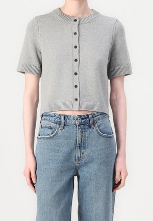 Mujer que lleva una chaqueta de punto corta de color gris claro con botones, mangas cortas, bordes acanalados y vaqueros azules de talle alto.