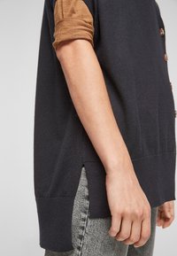 Mörk marinblå stickad topp med karamellfärgade uppvikta ärmar och bruna knappar, med sidospalter, matchad med grå texturerade byxor.