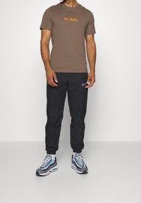 Hnědý tričko s nápisem "PARIS" v oranžové, spárované s černými tepláky s bílým logem Nike a šedobílými teniskami.