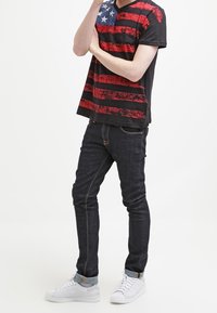 Svart T-shirt med röda ränder och blå stjärnmönster; ihopparad med mörka jeans och vita sneakers. Avslappnad design, standard passform.