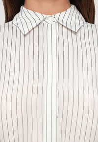Fehér blúz vékony fekete függőleges csíkokkal, hegyes gallérral és egyetlen látható gombbal a nyaknál.