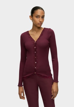 Kvinde med mørkt hår trukket tilbage iført en tætsiddende ribstrikket bordeaux cardigan med blondekant og knapper, kombineret med matchende bordeaux bukser.