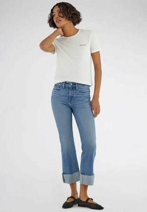 Donna che indossa una maglietta bianca a maniche corte con una piccola scritta, jeans azzurri con risvolto e ballerine nere chiuse, in piedi davanti a uno sfondo semplice.