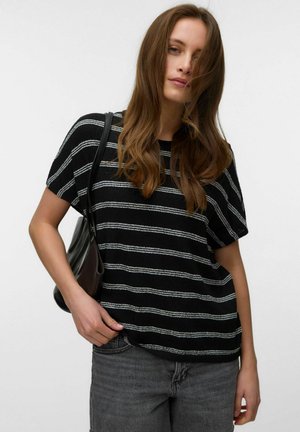 Donna con lunghi capelli castani che indossa una maglietta a righe bianco e nero a maniche corte, jeans grigi e porta una borsa nera a tracolla su sfondo bianco.