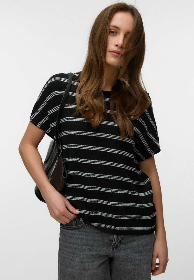 Femme aux longs cheveux bruns portant un haut à manches courtes rayé noir et blanc, un jean gris et un sac à main noir sur fond blanc.