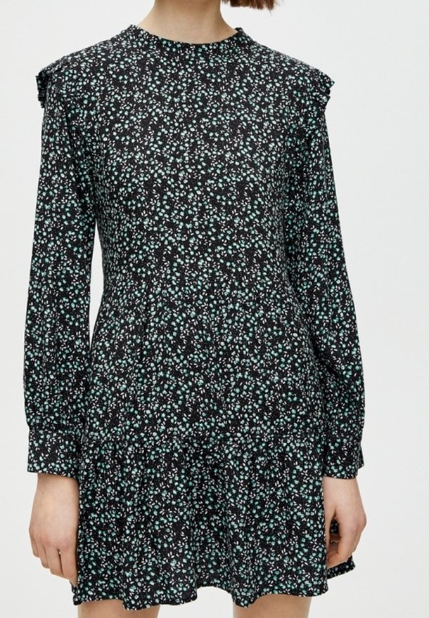 Femme portant une robe noire à manches longues avec un petit motif floral bleu clair et blanc et des détails à volants sur les épaules et l'ourlet.