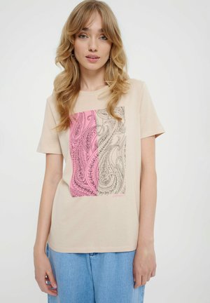 Młoda kobieta z blond falującymi włosami nosi beżowy T-shirt w różowe i beżowe wzory paisley oraz jasnoniebieskie dżinsy na białym tle.