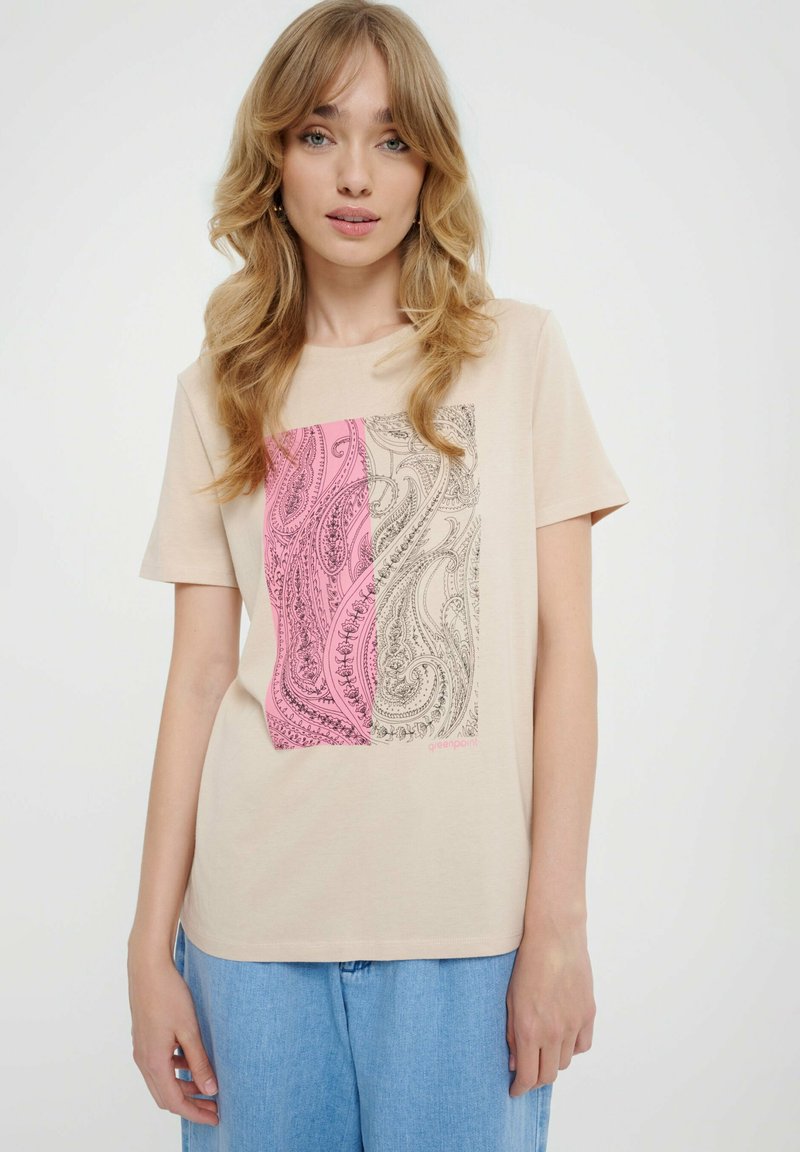 Młoda kobieta z blond falującymi włosami nosi beżowy T-shirt w różowe i beżowe wzory paisley oraz jasnoniebieskie dżinsy na białym tle.