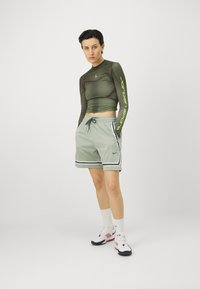Olívazöld, hosszú ujjú, crop top pöttyös mintával és zöld felirattal, amelyet világosszürke rövidnadrággal párosítottak, fehér oldalsávokkal és zsebekkel.