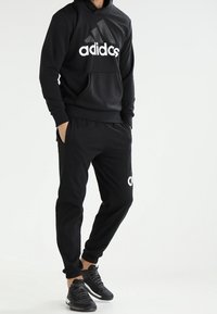 Uomo che indossa un hoodie nero Adidas, pantaloni jogger neri abbinati con logo e scarpe da ginnastica nere, in piedi con le mani in tasca contro uno sfondo bianco.