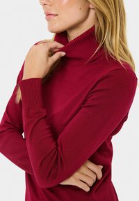 Burgundowy sweter z wysokim kołnierzem wykonany z gładkiego materiału, z prążkowanymi mankietami i kołnierzem, dopasowanym fasonem oraz długimi rękawami.