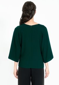 Blusa verde scuro con maniche a pipistrello ampie, scollatura rotonda e zip sul retro. Realizzata in tessuto morbido con una texture liscia.