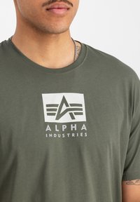 Olivově zelené tričko vyrobené z měkké tkaniny, které obsahuje bílé obdélníkové logo s textem "ALPHA INDUSTRIES" a stylizovaným designem "A".