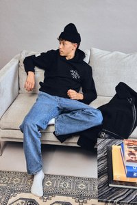 Jovem com um gorro preto e um hoodie reclinado num sofá bege, vestindo jeans azuis e meias brancas, ao lado de um casaco preto e livros empilhados na mesa.
