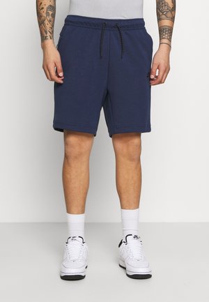 Mężczyzna ubrany w granatowe szorty na sznurek, białe skarpetki i białe trampki Nike Air Force 1, stojący na jasnym tle.