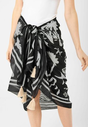 Paréo noir et gris à motifs avec des pompons beige noué autour de la taille sur un haut blanc, longueur moyenne couvrant les jambes jusqu'aux genoux.