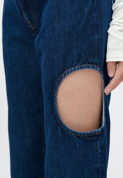 Vaqueros de mezclilla azul con un corte circular en la rodilla, con costuras naranjas en contraste y una textura suave. Un dedo toca el recorte.