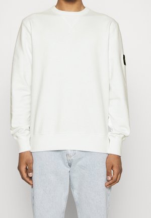 Man wearing plain white long-sleeve crewneck sweatshirt and light blue jeans, standing against a plain background.