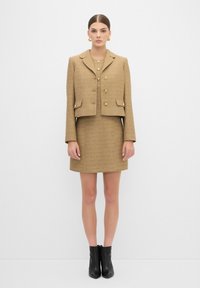Tan tweed-blazer med krage, fyra knappar och två framfickor, ihop med en matchande figursydd kjol. Svarta ankelskor fulländar looken.