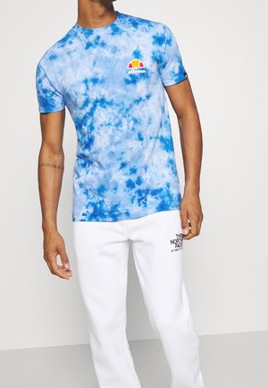 Blaues Batik-T-Shirt aus weichem Stoff, mit Rundhalsausschnitt und kleinem buntem Logo auf der Brust. Kombiniert mit weißen Jogginghosen.