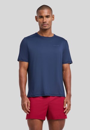 Mann trägt ein marineblaues Kurzarmhemd und rote Sportshorts und steht vor einem einfarbigen grauen Hintergrund.