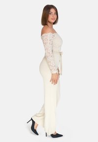 Top in pizzo scivolato sulle spalle di colore crema abbinato a pantaloni ampi crema. Tacchi a spillo neri aggiungono contrasto. La modella è in profilo.