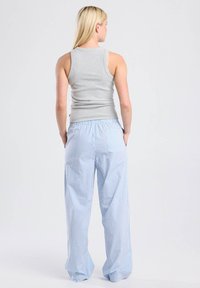 Mujer con cabello largo rubio, vestida con una blusa sin mangas gris y pantalones sueltos a rayas azul y blanco, de pie con las manos en los bolsillos, vista por detrás.