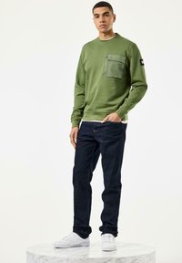 Grüner Sweatshirt mit einer Reißverschlusstasche auf der Brust, kombiniert mit dunkelblauen Jeans und weißen Sneakern, vor einem neutralen Hintergrund.