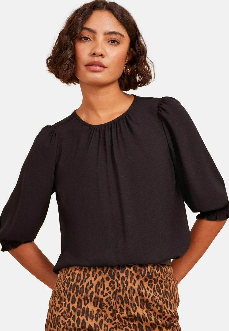 Schwarze Bluse mit kurzen Ärmeln und geraffter Textur an den Schultern und am Hals. Kombiniert mit gemusterten Leoparden-Hosen.