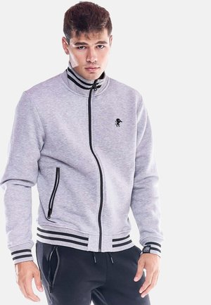Giovane uomo con una giacca leggera grigio chiaro con zip, con strisce nere sul colletto, polsini e orlo, abbinata a pantaloni neri su uno sfondo semplice.