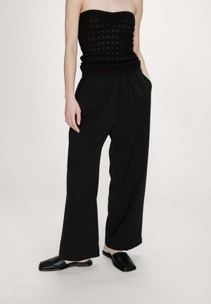 Femme portant un top en maille noir sans bretelles, un pantalon noir à jambes larges avec les mains dans les poches, et des chaussures plates noires à enfiler sur un fond uni.