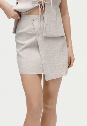 Beige linen wrap skirt with two buttons and asymmetrical hem worn with matching top showing tied strings and bare legs.