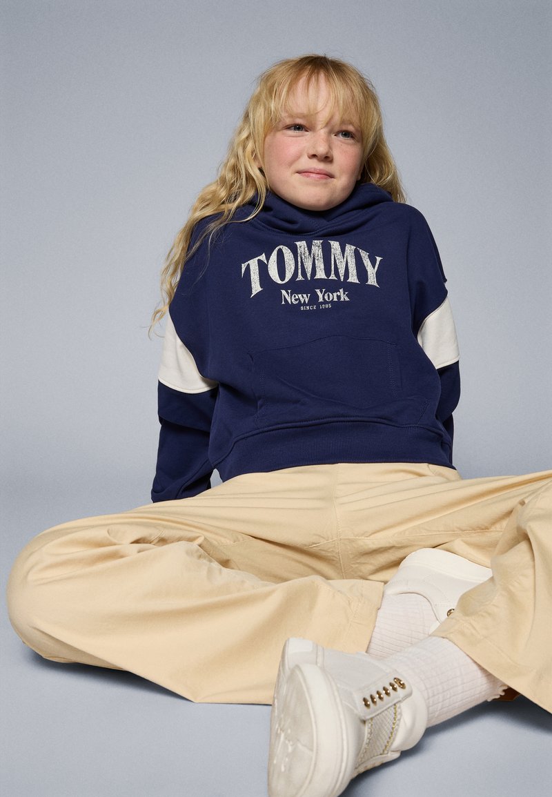 Marineblå hettegenser med hvite ermer, "Tommy New York" tekst. Beige vide bukser, hvite sneakers og teksturerte hvite sokker.