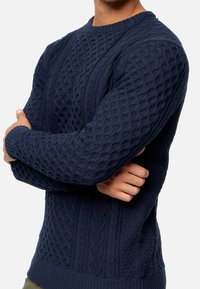 Navy blue knitted sweater featuring textured patterns, ribbed cuffs and hem, and a crew neck design. Made from a thick, cozy fabric.