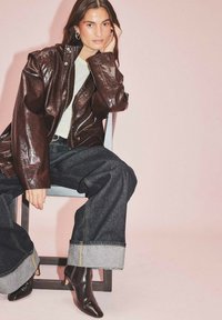 Rjav usnjen jopič, svetla majica in široke temne kavbojke z zavihanimi robovi. Poudarjeno z črnimi čevlji na visoki peti. Enostavna kulisa.