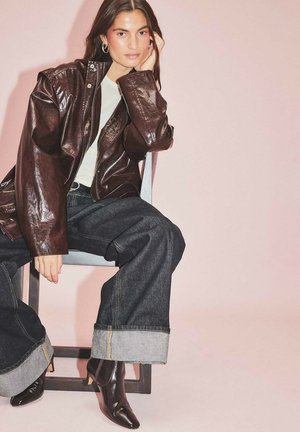 Chaqueta de cuero marrón, parte superior clara y jeans de mezclilla oscuros de pierna ancha con dobladillo enrollado. Acento con botas de tobillo negras de tacón. Fondo sencillo.
