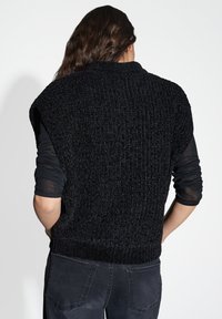 Maglione senza maniche nero lavorato a maglia con un motivo testurizzato e orlo a coste, indossato sopra una camicia nera a maniche lunghe, abbinato a jeans scuri.