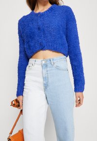 Žena v krátkém modrém chlupatém svetru s knoflíky a dvoubarevných džínech s vysokým pasem, držící oranžovou kabelku za popruh.