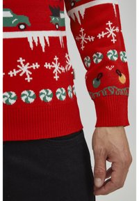 Roter gestrickter Pullover mit grünen Autos und weißen Schneeflocken. Enthält Zuckerstangenmuster und bunte Ornamente am Saum.