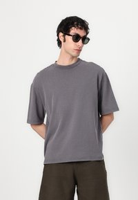 T-shirt gris à manches courtes avec un col rond, coupe légèrement ample et coutures visibles. Associé à un short vert foncé.