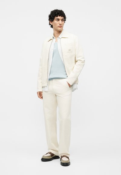 Άνδρας που φορά κρεμ σακάκι, παντελόνι στο ίδιο χρώμα, ανοιχτό μπλε ριπ πλεκτό γιλέκο, λευκό πουκάμισο και καφέ-κρεμ μοκασίνια, στέκεται με το ένα χέρι στην τσέπη.