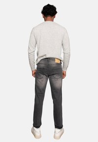 Jeans gris para hombre con diseño desgastado, estilo de cinco bolsillos y una etiqueta de cuero marrón en la parte posterior de la cinturilla. Combinados con una camiseta de manga larga gris claro.