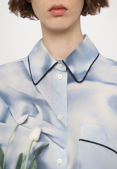 Light blue blouse in sheer fabric, featuring a flowing design, black piping, and white floral print. Includes a pocket with accent stitching.